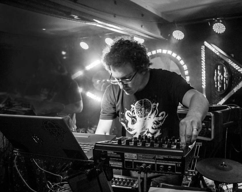 Ed Guild in a dim club, leaning over a mixer and laptop, adjusting a knob